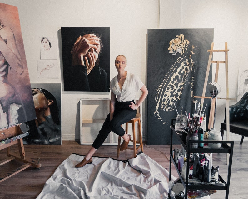 May include: A woman wearing a white shirt and black pants sits on a stool in an art studio. She is surrounded by paintings, including a portrait of a woman with her hands covering her face, a cheetah, and a nude figure. The studio is well-lit and has a wooden floor.
