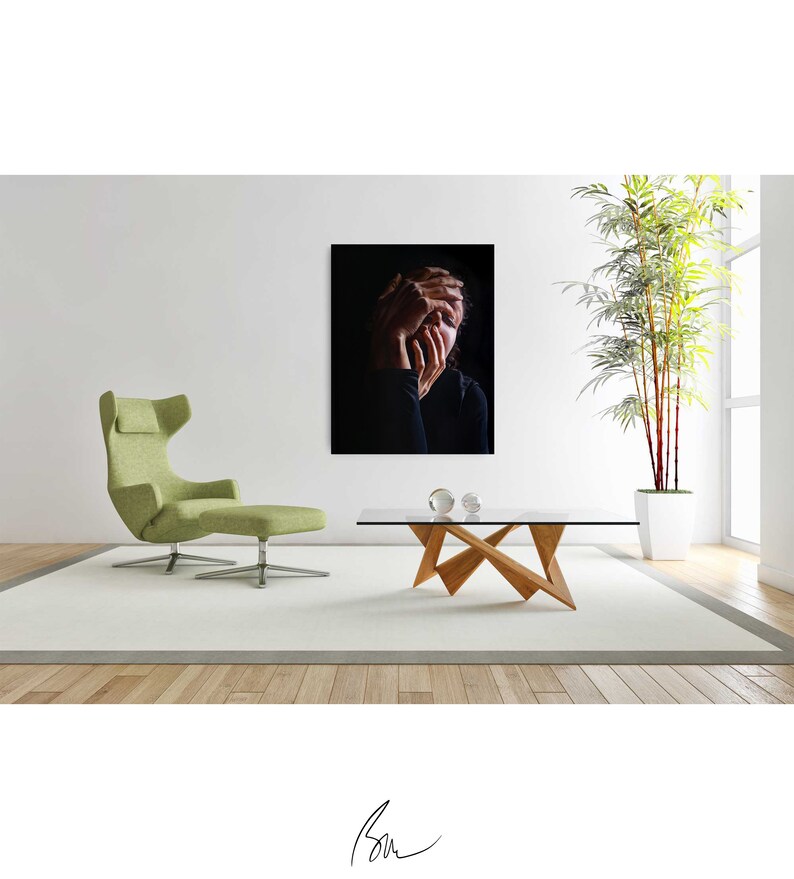 May include: A black and white photograph of a person with their hands covering their face. The photo is framed and hanging on a white wall above a wooden coffee table.