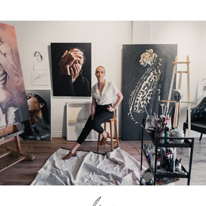 Op de afbeelding: Een vrouw in een wit blouse en zwarte broek zit in een kunststudio. Ze is omringd door schilderijen, waaronder een portret van een vrouw met haar handen voor haar gezicht, een cheetah en een portret van een vrouw met haar hoofd naar achteren gekanteld. De studio is ingericht met een houten ezel, een zwarte metalen kar met kunstbenodigdheden en een zwarte leren stoel.