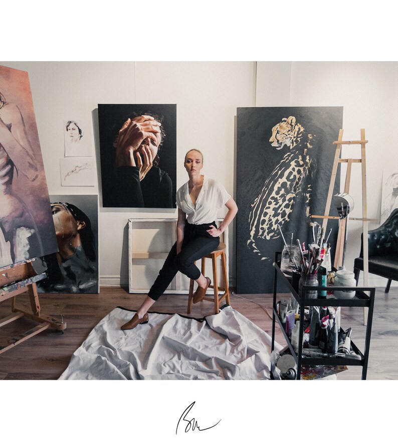 Op de afbeelding: Een vrouw in een wit blouse en zwarte broek zit in een kunststudio. Ze is omringd door schilderijen, waaronder een portret van een vrouw met haar handen voor haar gezicht, een cheetah en een portret van een vrouw met haar hoofd naar achteren gekanteld. De studio is ingericht met een houten ezel, een zwarte metalen kar met kunstbenodigdheden en een zwarte leren stoel.