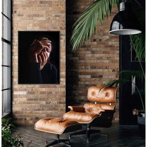 May include: A black and white photo of a person with their hands covering their face. The photo is framed and hanging on a brick wall. A brown leather armchair and ottoman are in the foreground.
