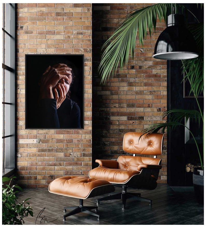 May include: A black and white photo of a person with their hands covering their face. The photo is framed and hanging on a brick wall. A brown leather armchair and ottoman are in the foreground.