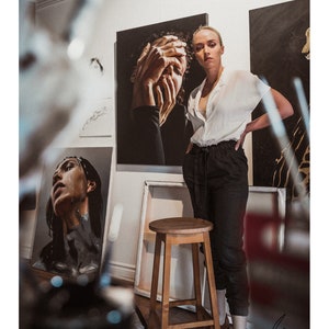 May include: A woman stands in an art studio, wearing a white button-down shirt and black pants. She is standing in front of a painting of a woman with her hands covering her face. There is a wooden stool in the foreground.