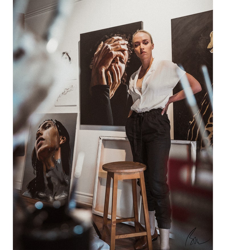 May include: A woman stands in an art studio, wearing a white button-down shirt and black pants. She is standing in front of a painting of a woman with her hands covering her face. There is a wooden stool in the foreground.