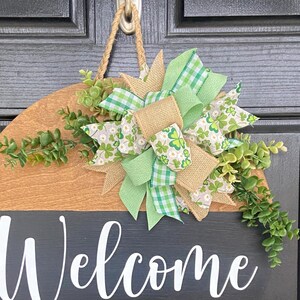 May include: A round wooden welcome sign with a black and brown color scheme. The word "Welcome" is written in white script. The sign is decorated with green and white bows and greenery, and is hanging on a black door.