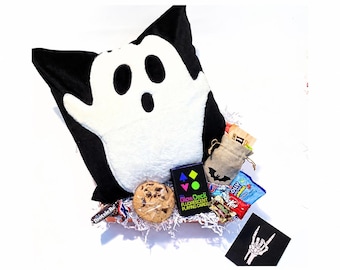 May include: A gift basket containing a black and white ghost-shaped plush pillow, a deck of GlowDeck fluorescent playing cards, a chocolate chip cookie, various candy bars, a small burlap drawstring bag with a bat icon, and a black card with a skeleton hand.