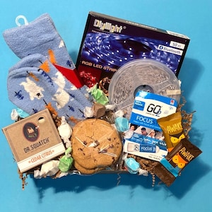 May include: A gift basket filled with various items, including a pair of blue socks with a white pattern, a roll of white LED light strip, a brown bar of soap, a package of chocolate bars, and two packages of chocolate-covered cookies.