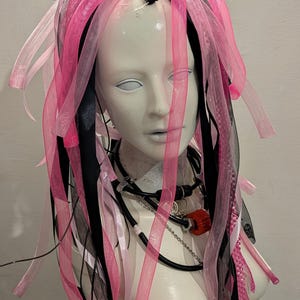 Könnte beinhalten: Ein Schaufensterpuppenkopf mit rosa, schwarzen und weißen Bändern und Stoffsträngen, die als Haare gestylt sind. Die Schaufensterpuppe trägt eine Halskette mit rotem Anhänger. Die Büste ist weiß und glatt.