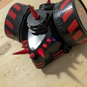 以下が含まれることがあります： 赤と黒のスパイクとストライプが特徴の黒いガスマスク。マスクには、赤と黒のデザインの2つの円形フィルターがあります。全体的なデザインはエッジの効いたインダストリアルです。