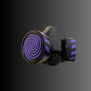 Pu&ograve; includere: Una maschera antigas nera con un design a spirale viola e nero sul davanti. La maschera ha un cinturino nero e un filtro nero.