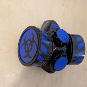 Op de afbeelding: Zwart en blauw ademhalingsmasker met een biohazard-symbool. Het masker heeft blauwe accenten en verstelbare knoppen. Ontworpen voor luchtfiltratie en bescherming van de drager.