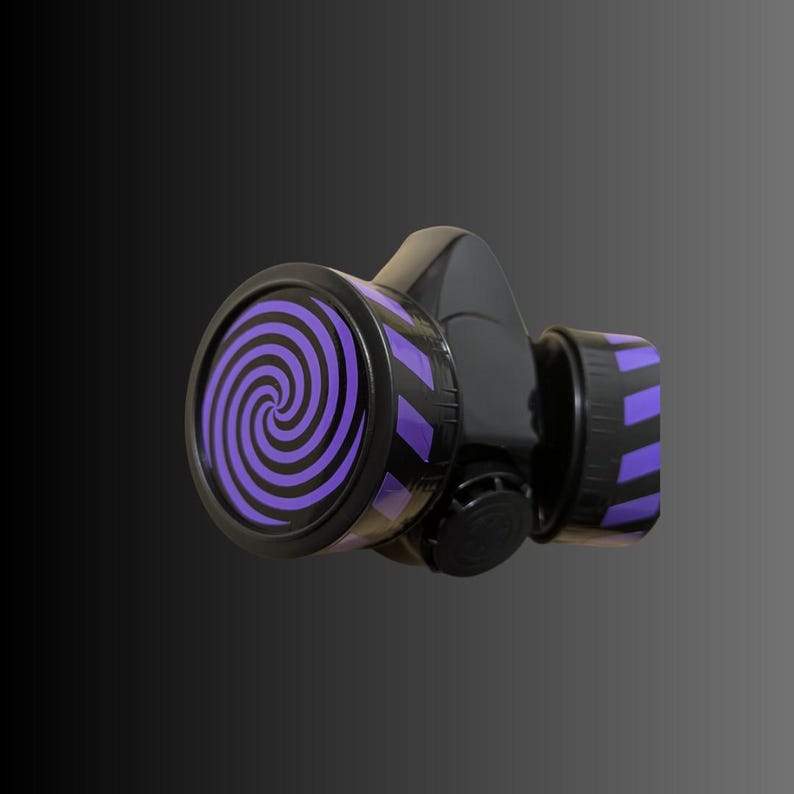 Pu&ograve; includere: Una maschera antigas nera con un design a spirale viola e nero sul davanti. La maschera ha un cinturino nero e un filtro nero.