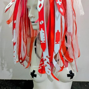 Könnte beinhalten: Ein Mannequin-Torso mit einem medizinisch thematisierten Kopfschmuck. Der Kopfschmuck zeigt rote und weiße Bänder, medizinische Symbole und Spritzenmotive. Der Torso hat schwarze, kreuzförmige Brustwarzenabdeckungen.