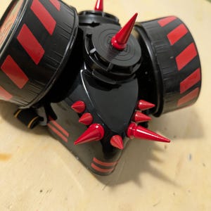 以下が含まれることがあります： 赤と黒のアクセントとスパイクが付いた黒い呼吸マスク。マスクには、赤い斜めのストライプと複数の赤いスパイクが付いた2つの大きな円形フィルターがあります。マスクは光沢のある仕上げと詳細なデザインで設計されています。