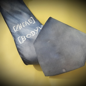 Könnte beinhalten: Eine blaue Krawatte mit den Worten "HEAD" und "BODY" in weißem Text. Die Krawatte hat einen Farbverlauf. Die Krawatte liegt auf einer gelben Oberfläche.