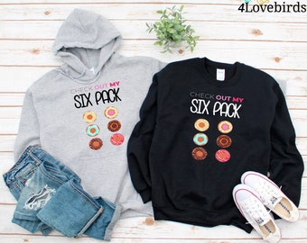 May include: A gray hoodie and a black sweatshirt are displayed, both featuring the text "CHECK OUT MY SIX PACK" above a graphic of six donuts. The donuts have various colors and toppings. Blue jeans and white sneakers are also visible.