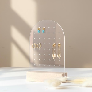 May include: Clear acrylic earring display stand with a wooden base. The stand features multiple holes for hanging earrings. Several pairs of earrings are displayed, including gold hoops, turquoise studs, and dangling pearl earrings. The stand is arched at the top.