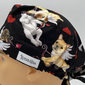 Könnte beinhalten: Schwarze OP-Mütze mit einem Aufdruck von Hunden, die als Amor mit Flügeln und Pfeilen verkleidet sind. Die Mütze hat einen schwarzen Kordelzug und ein weißes Etikett mit der Aufschrift "Nuestra Vida".