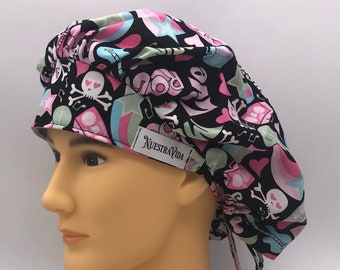 May include: A black scrub hat with a colorful print featuring skulls, hearts, stars, and musical notes. The hat has a tie at the back and a label that reads "Nuestra Vida".
