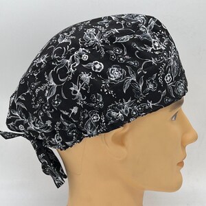 Könnte beinhalten: Schwarze OP-Mütze mit weißem Blumenmuster. Die Mütze hat einen Bindegurt an der Rückseite.