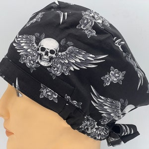 Könnte beinhalten: Schwarze OP-Mütze mit Totenkopf- und Flügelmotiv. Der Totenkopf ist von Rosen umgeben und die Flügel sind ausgebreitet. Die Mütze hat einen Bindegurt an der Rückseite.