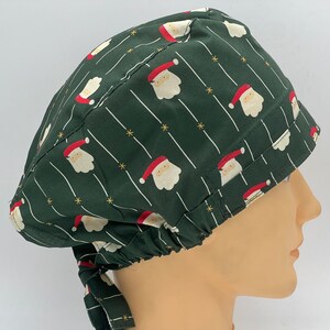 Könnte beinhalten: Grüne OP-Mütze mit einem sich wiederholenden Muster aus Weihnachtsmann-Gesichtern und goldenen Sternen. Die Mütze hat einen Bindfaden an der Rückseite.