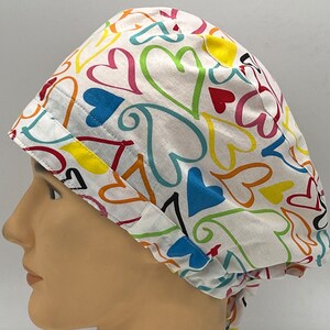 Könnte beinhalten: Eine weiße OP-Mütze mit einem bunten Druck von Herzen in verschiedenen Schattierungen von Rosa, Blau, Grün, Gelb und Orange. Die Herzen sind in einem verspielten Stil gezeichnet.