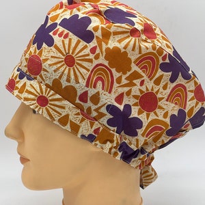 Könnte beinhalten: Eine weiße OP-Mütze mit einem bunten Muster aus Sonnen, Regenbögen und Wolken in Orange-, Rot-, Lila- und Brauntönen.