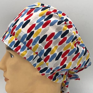 Könnte beinhalten: Eine weiße OP-Mütze mit einem bunten geometrischen Muster in Rot-, Blau-, Gelb- und Schwarztönen. Die Mütze hat einen Bindfaden an der Rückseite.
