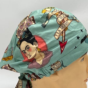 Könnte beinhalten: Eine türkisblaue OP-Mütze mit einem farbenfrohen Tattoo-inspirierten Druck, der Frida Kahlo, Blumen und einen Totenkopf mit dem Text "YIA MUERTE E PLATE" zeigt.