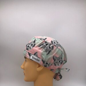 Könnte beinhalten: Eine rosa und grüne OP-Mütze mit einem sich wiederholenden Muster aus schwarzen und weißen Katzenköpfen. Die Mütze hat einen Bindegurt am Rücken und ein kleines weißes Etikett mit dem Text "Nuestra Vida".