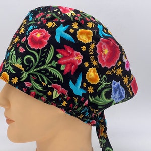 Könnte beinhalten: Eine schwarze OP-Mütze mit einem bunten Blumen- und Vogelmuster. Die Mütze hat einen Bindegurt an der Rückseite.
