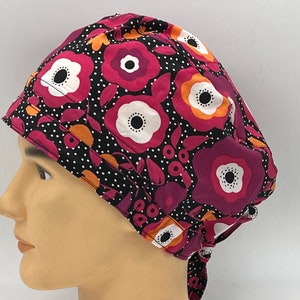Könnte beinhalten: Eine schwarze OP-Mütze mit einem Blumenmuster aus rosa, orangefarbenen und weißen Blüten auf schwarzem Grund mit weißen Punkten. Die Mütze hat einen Bindegurt am Rücken.