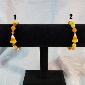 Può includere: Due braccialetti a tema mais dolce su un espositore nero. I braccialetti sono realizzati con perline arancioni, gialle e bianche. I braccialetti sono etichettati con il numero 1 e 2.