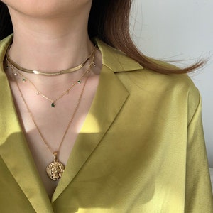 Op de afbeelding: Een gelaagde gouden ketting met een muntbedel en groene edelsteenaccenten. De ketting wordt gedragen door een persoon met lang bruin haar en een geel shirt.