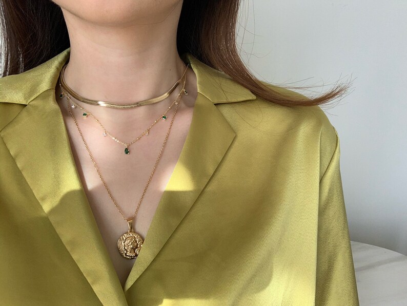 Op de afbeelding: Een gelaagde gouden ketting met een muntbedel en groene edelsteenaccenten. De ketting wordt gedragen door een persoon met lang bruin haar en een geel shirt.