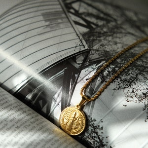 Op de afbeelding: Een gouden ketting met een ronde hanger met een religieus beeld. De ketting ligt op een boek met een zwart-witfoto van een gebouw en bomen.