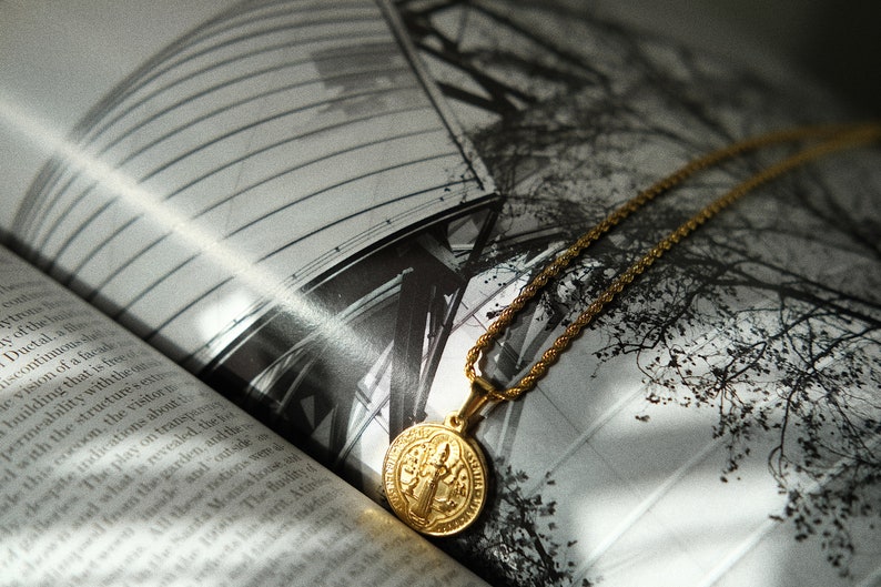 Op de afbeelding: Een gouden ketting met een ronde hanger met een religieus beeld. De ketting ligt op een boek met een zwart-witfoto van een gebouw en bomen.