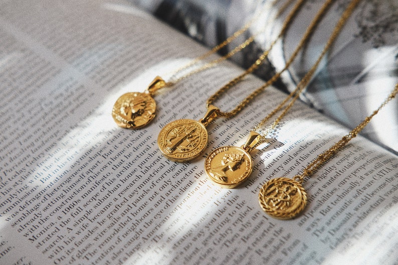 Op de afbeelding: Vier gouden kettingen met munt hangers met verschillende ontwerpen. De kettingen liggen op een boek met tekst.