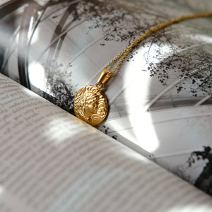 Op de afbeelding: Een gouden ketting met een ronde hanger met een gedetailleerd profiel van een vrouwenhoofd. De ketting ligt op een boek met een zwart-witfoto van een gebouw.