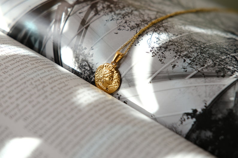 Op de afbeelding: Een gouden ketting met een ronde hanger met een gedetailleerd profiel van een vrouwenhoofd. De ketting ligt op een boek met een zwart-witfoto van een gebouw.