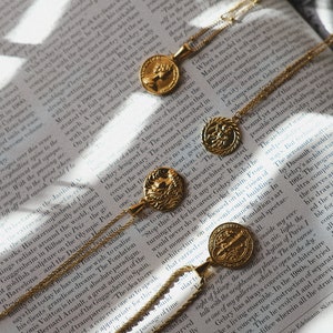 Op de afbeelding: Drie gouden kettingen met muntpendanten met ingewikkelde ontwerpen. De munten zijn in verschillende maten en vormen, sommige met reli&euml;fdetails. De kettingen liggen op een boek met tekst.