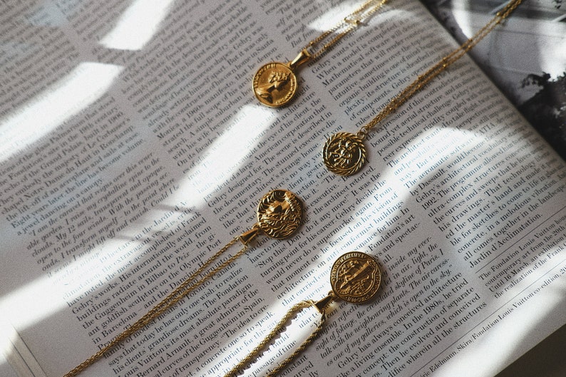 Op de afbeelding: Drie gouden kettingen met muntpendanten met ingewikkelde ontwerpen. De munten zijn in verschillende maten en vormen, sommige met reli&euml;fdetails. De kettingen liggen op een boek met tekst.