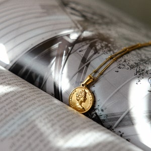 Op de afbeelding: Een gouden ketting met een ronde hanger met een profiel van een vrouwenhoofd. De ketting ligt op een boek met een zwart-witfoto van een gebouw.