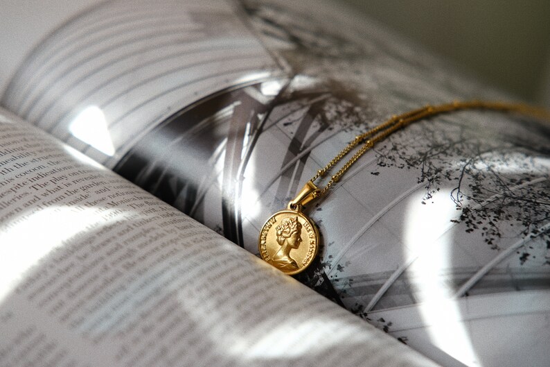 Op de afbeelding: Een gouden ketting met een ronde hanger met een profiel van een vrouwenhoofd. De ketting ligt op een boek met een zwart-witfoto van een gebouw.