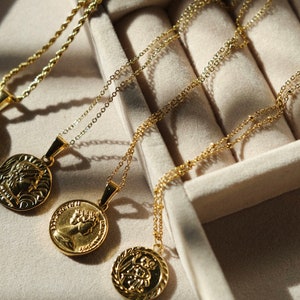 May include: Four gold necklaces with different coin-shaped pendants in a jewelry box. The pendants feature various designs, including a profile of a person, a religious figure, and a floral motif.