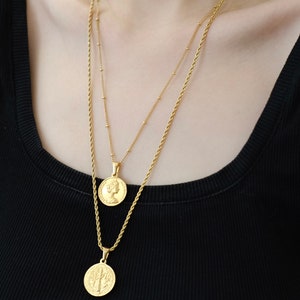 Op de afbeelding: Twee gouden kettingen met munt hangers. De bovenste ketting heeft een kleine munt met het profiel van een vrouw. De onderste ketting heeft een grotere munt met een boomontwerp.