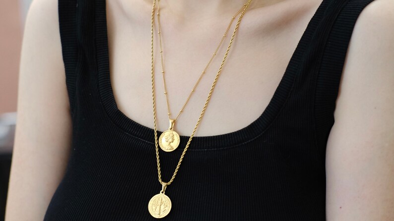 Op de afbeelding: Twee gouden kettingen met munt hangers. De bovenste ketting heeft een kleine munt met het profiel van een vrouw. De onderste ketting heeft een grotere munt met een boomontwerp.