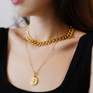 Op de afbeelding: Een vrouw draagt een zwart tanktop met twee gouden kettingen. De bovenste ketting is een dikke, stoere ketting. De onderste ketting is een delicate ketting met een gouden munt hanger.