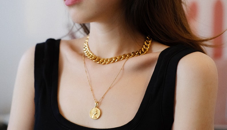 Op de afbeelding: Een vrouw draagt een zwart tanktop met twee gouden kettingen. De bovenste ketting is een dikke, stoere ketting. De onderste ketting is een delicate ketting met een gouden munt hanger.